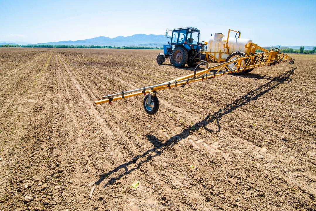 Синий трактор с желтым опрыскивателем на пахотном поле под ясным небом, на фоне гор.
