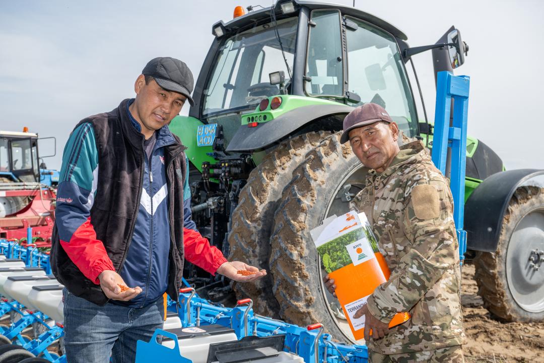 Два фермера с семенами и упаковкой на фоне трактора и сеялки в поле.