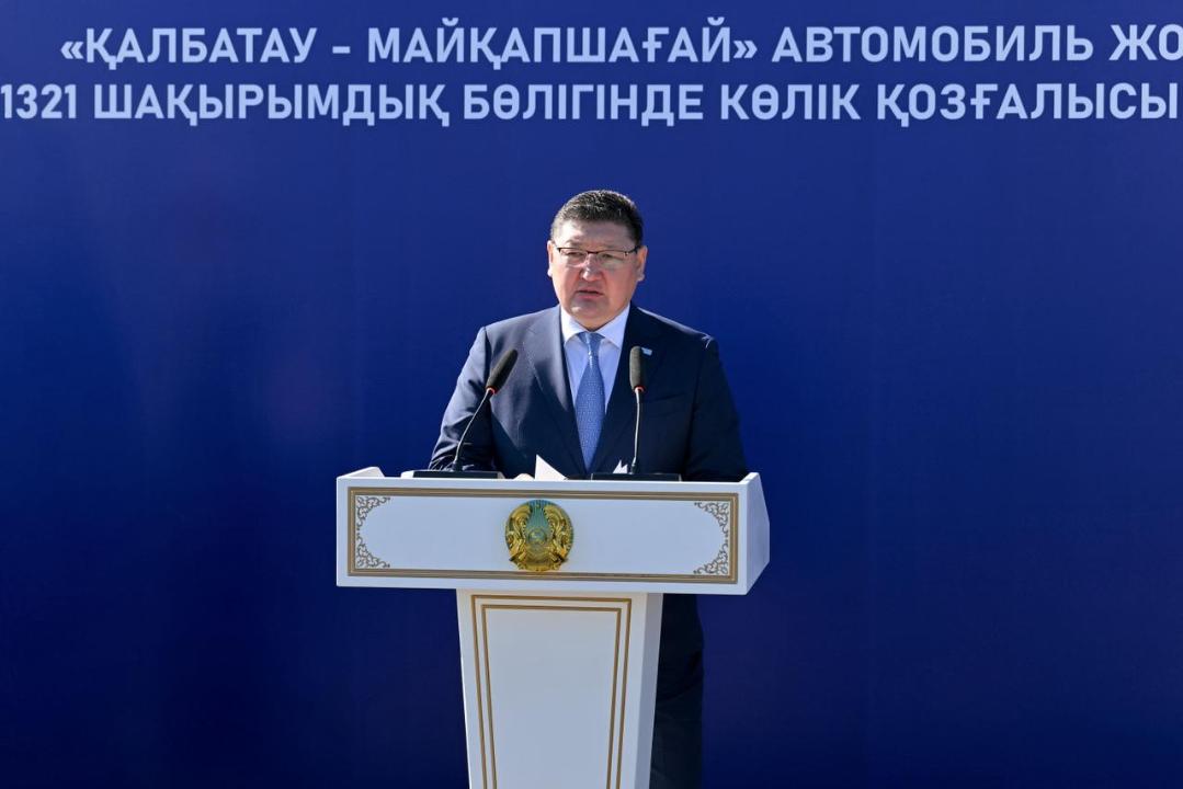 Бұл фотосуретте «Қалбатау – Майқапшағай» жолына арналған баннердің алдында трибунада сөз сөйлеп тұр