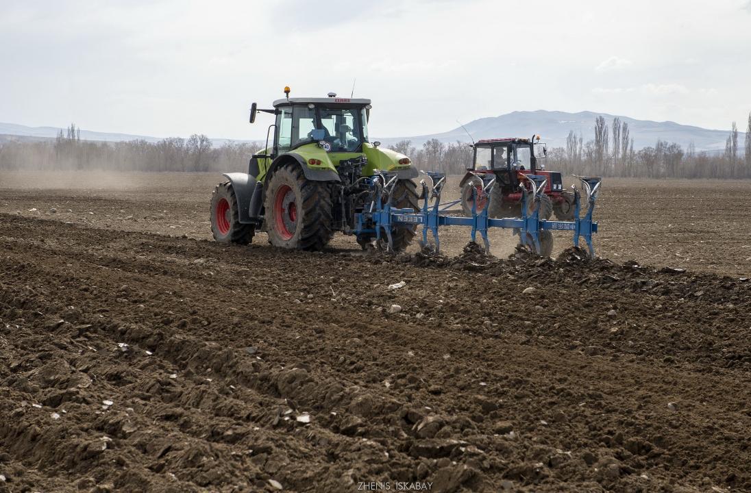 Зелёный трактор с плугом пашет поле, за ним следует красный трактор. На заднем плане &mdash; деревья и горы.