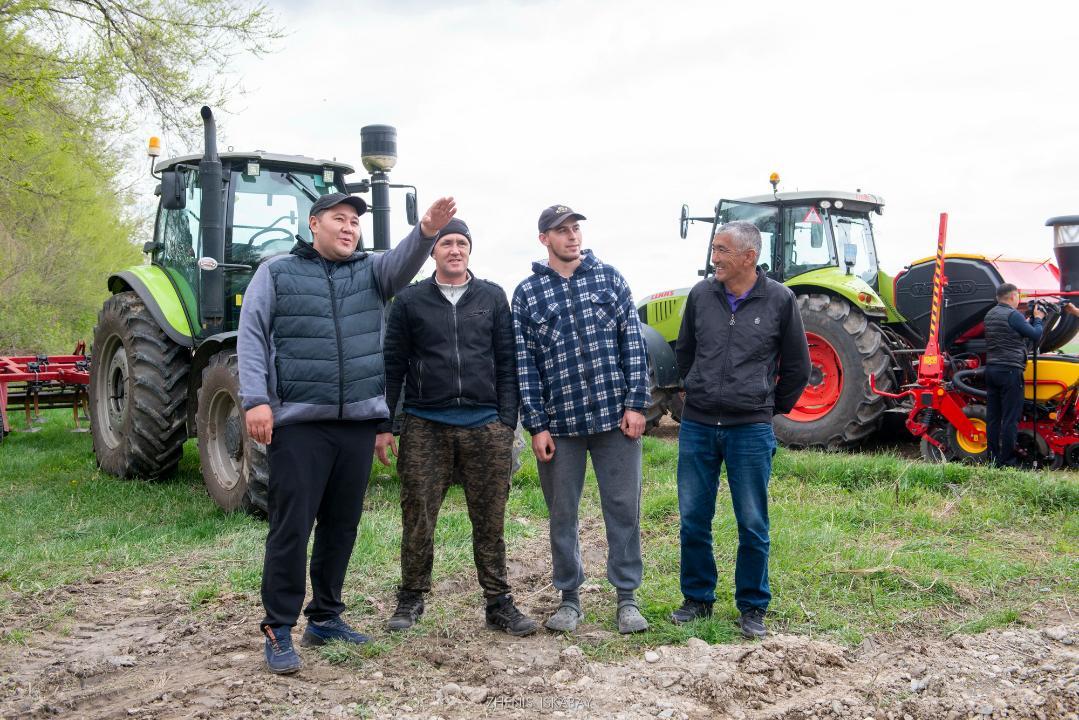 Четыре мужчины стоят на поле перед зелеными тракторами, один указывает вдаль рукой.