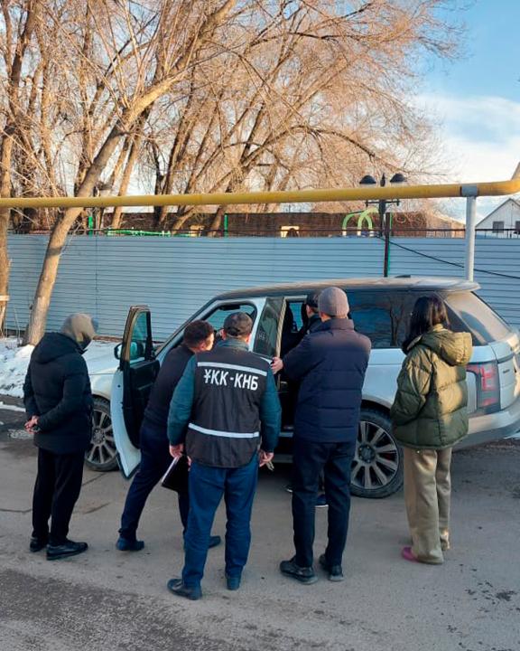 Сотрудники УКК-КНБ в жилетах осматривают белый внедорожник на улице зимой, рядом стоят гражданские.